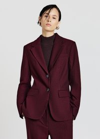 Blazer bordeaux con righe sottili, due bottoni, collo a sciarpa e tasche a pattina. Realizzato in tessuto testurizzato, abbinato a un top in maglia scura.