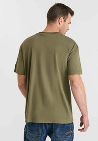 Camiseta de manga corta color verde oliva hecha de tela suave. Presenta un escote redondeado y un corte relajado, con costuras visibles en la parte posterior.