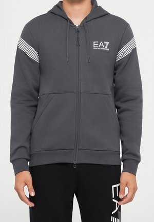 Mand iført en mørkegrå hættetrøje med lynlås med hvide striber på ærmerne og EA7 Emporio Armani-logo på brystet.