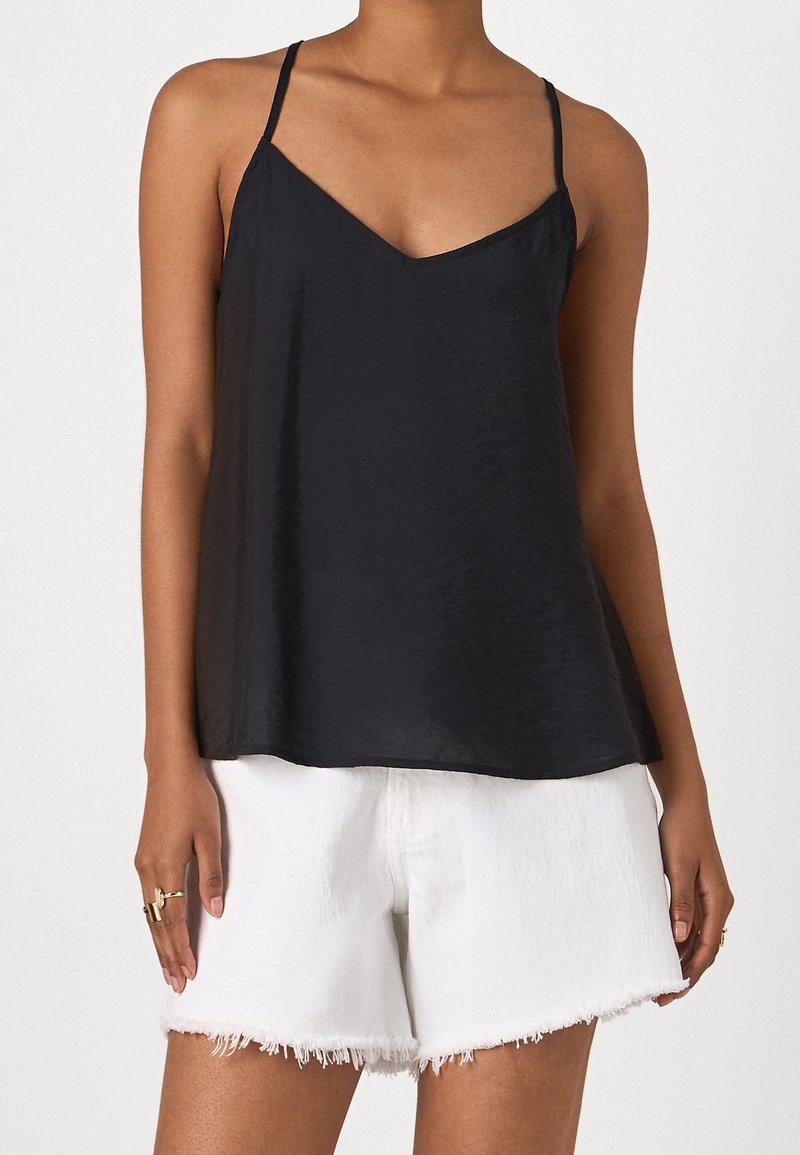 Femme portant un caraco noir sans manches à col en V et un short en jean blanc effiloché, avec des bagues aux doigts, sur un fond uni.