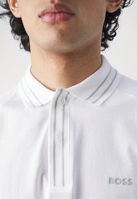 Polo blanc avec patte à deux boutons, col texturé avec des rayures grises et un logo subtil "BOSS" sur la poitrine gauche.