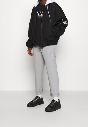Man wearing black bomber jacket with patch, black graphic sweatshirt, light gray cargo pants, white socks, and black sneakers standing on white floor.
