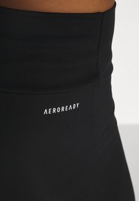 Czarne legginsy o wysokim stanie, wykonane z elastycznej tkaniny, z subtelną teksturą i białym logo "AEROREADY" z przodu.