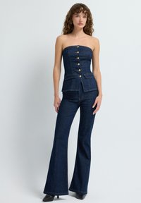 Džínový top bez ramínek se zlatými knoflíky, přiléhavý korzet, kalhoty s rozšířenými nohavicemi a kontrastní švy, vše v tmavě modré barvě. Hladká textura a na míru šitý design.