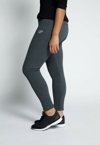 Donkergrijze leggings van rekbaar materiaal met een gladde textuur, met een logo "UP" op de heup en een strakke pasvorm. Zwarte sportschoenen zijn gedragen.