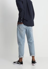 Jasnoniebieskie dżinsy z podwiniętymi nogawkami, w połączeniu z czarnymi sneakersami z białymi podeszwami. Model nosi ciemną koszulę z guzikami.