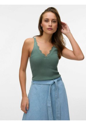 Ženska, oblečena v zeleno rebrasto pleteno majico brez rokavov z valovitim dekoltejem in svetlo modro denim krilo z visokim pasom ter pasom, ki je privezan.