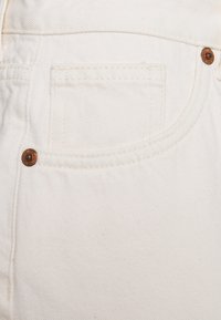Primer plano de tela de mezclilla blanca con costuras detalladas, remaches de cobre y un pequeño bolsillo en la parte frontal de los vaqueros.