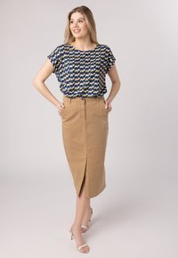 Printblouse met marineblauw, geel en witte patronen, gedragen bij een beige rok tot de knie met een voorste split en knoopsluiting.