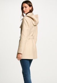 Beige trenchcoat met een capuchon, een ceintuur in de taille en brede schouders. Gemaakt van een gladde stof; heeft een middellange snit en zijzakken.
