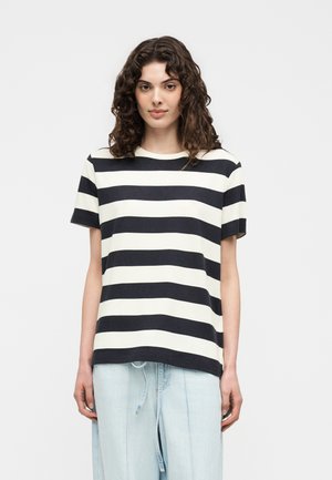 Vrouw met krullend bruin haar, die een zwart-wit breed horizontaal gestreept T-shirt draagt en lichtblauwe losse broek tegen een effen achtergrond.