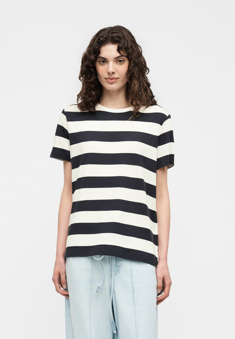 Γυναίκα με καστανά κατσαρά μαλλιά που φοράει μαύρο και άσπρο φαρδύ οριζόντιο ριγέ μπλουζάκι και ανοιχτό μπλε φαρδιά παντελόνια σε απλό φόντο.