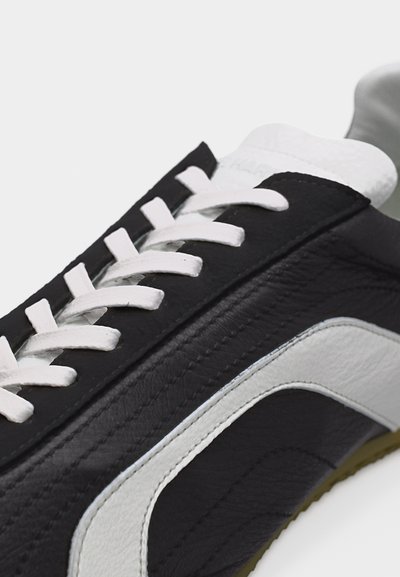 Ασπρόμαυρο δερμάτινο αθλητικό παπούτσι με λευκά κορδόνια και μια καμπυλωτή λευκή ρίγα στο πλάι, σε κοντινή λήψη σε ανοιχτό φόντο.