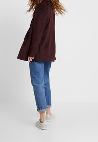 Blazer bordeaux avec une texture douce, un design ajusté et des boutons noirs, associé à un jean en denim bleu retroussé et des baskets gris clair.