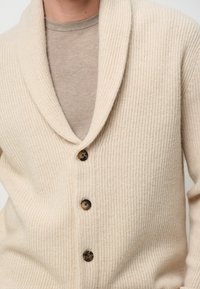 Beige gestrickte Strickjacke mit Schalkragen, ausgestattet mit drei großen braunen Knöpfen und einem strukturierten Rippenmuster, über einem hellbeigen Hemd.
