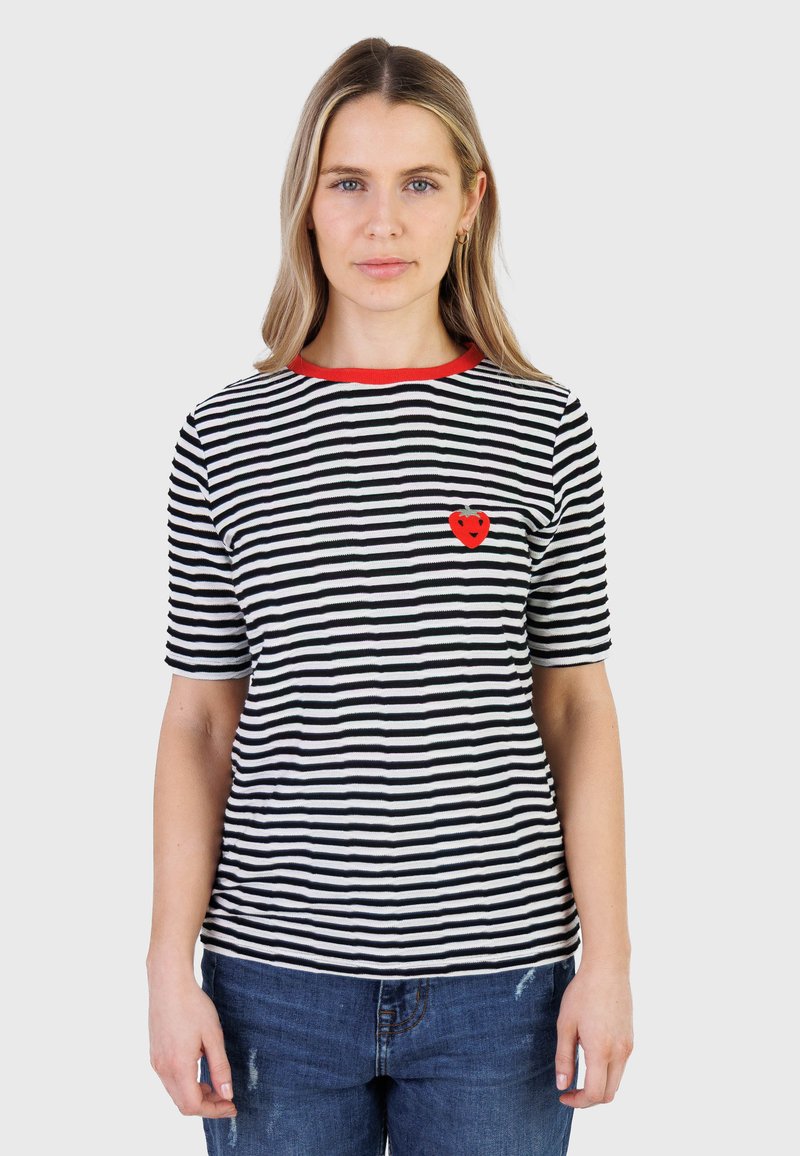 Mujer con cabello rubio que lleva una camiseta de rayas blancas y negras con un emblema de corazón rojo y cuello rojo, combinada con jeans azules.