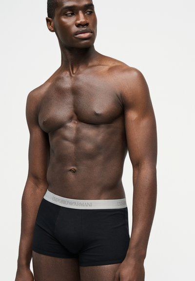 Särgita mees, kellel on lihaseline kehaehitus, kannab musti Emporio Armani bokserpükse ja vaatab vasakule tavalise heleda tausta taustal.