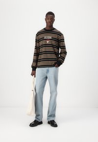 Langermet svart stripete skjorte med "GUESS"-logo, blå denimjeans, svarte sko og en beige totebag. Enkel, uformell design.