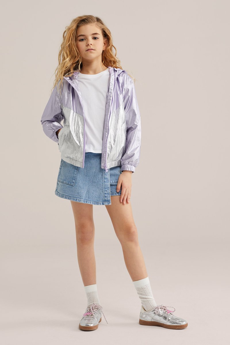 Mlada deklica z valovitimi blond lasmi, oblečena v sijočo vijolično-srebrno jakno, belo majico, modro denim krilo, bele nogavice in srebrne čevlje.