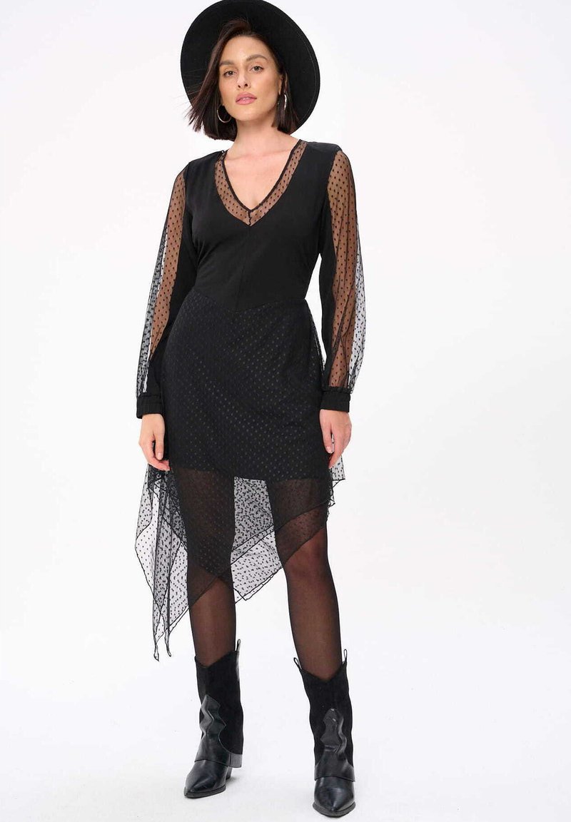 Zwarte jurk met doorzichtige, stippenmouwen en een asymmetrische onderkant. Heeft een diepe V-hals en een gelaagde textuur. Gecombineerd met zwarte laarzen.