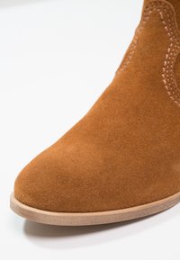Gros plan sur l'avant d'une botte en daim marron avec des coutures détaillées et une semelle en bois clair sur fond blanc.