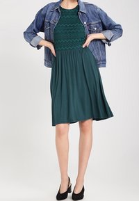 Robe verte avec un corsage texturé et une jupe lisse, associée à une veste en denim. Le mannequin porte des chaussures noires à bouts fermés.