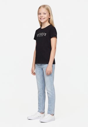 Nuori tyttö seisoo hymyillen, yllä musta Tommy Hilfiger -t-paita, vaaleansiniset farkut ja valkoiset lenkkarit valkoista taustaa vasten.