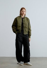 Βερμουδαράκι bomber σε ελιά πράσινο με φερμουάρ, πλαϊνές τσέπες, συνδυασμένο με μαύρο cargo παντελόνι. Το μοντέλο φοράει γκρι αθλητικά παπούτσια. Απλός, σύγχρονος σχεδιασμός.