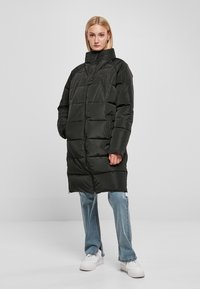Cappotto lungo nero imbottito con colletto alto e chiusura a zip. Tessuto liscio, tasche laterali e vestibilità ampia, indossato sopra jeans blu e sneakers bianche.