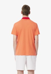 Polo a maniche corte di un brillante arancione con colletto rosso, caratterizzato da un logo discreto sul retro, realizzato in tessuto liscio e traspirante.
