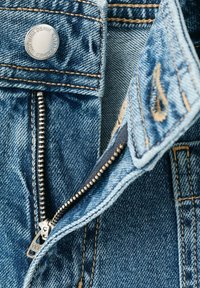 Blugi albaștri din denim despătușiți, arătând fermoarul metalic, nasturele cu textul "Denim Established 1984" și detalii de cusături portocalii.