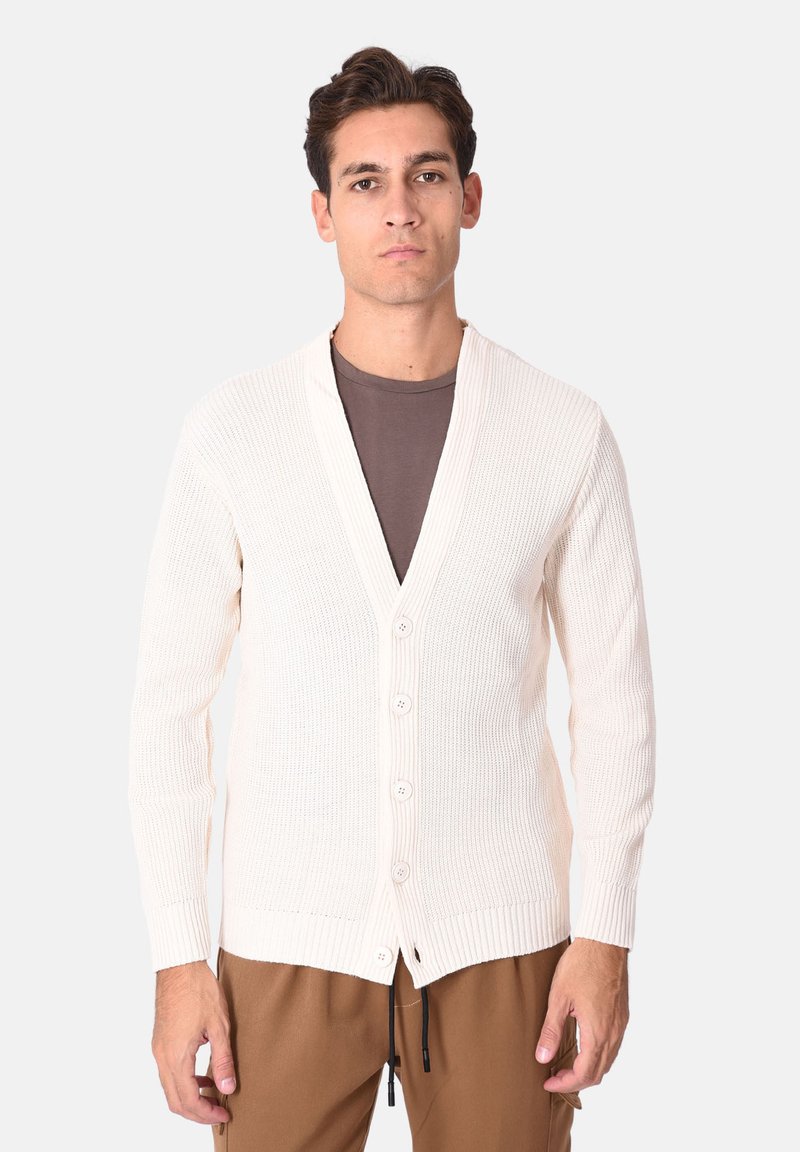 Cardigan lavorato a maglia color crema con scollo a V, cinque bottoni e polsini a costine. Tessuto morbido, taglio ampio e maniche a lunghezza media. Abbinato a pantaloni marroni.