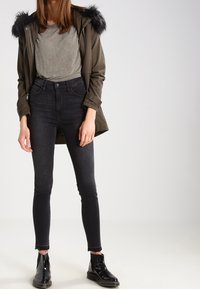 Žena v olivově zelené parce s kožešinovým lemem, šedém tričku, černých skinny džínách a černých lakovaných kozačkách.