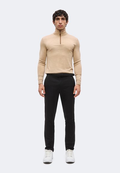 Homme debout portant un pull à manches longues beige avec fermeture éclair, pantalon noir avec ceinture et baskets blanches sur un fond clair uni.