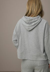 Sivý skrátený sweatshirt s kapucňou, vyrobený z textúrovanej látky a s rebrovanými detailmi, nosený s menej výraznými sivými nohavicami. Zobrazený zo zadnej strany.