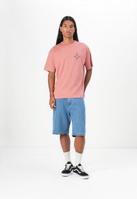 Light pink cotton t-shirt with a graphic design on the chest, paired with blue denim shorts, white crew socks, and black sneakers.