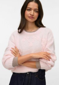 Jasnoróżowy dziergany sweter z motywem aplikacji kwiatowej, szare prążkowane mankiety, luźny krój, noszony z ciemnymi dżinsami z detalem róży.