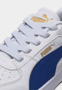 Λευκό αθλητικό παπούτσι Puma με μπλε πλευρική ρίγα, λευκά κορδόνια και χρυσά λογότυπα Puma στη γλώσσα και την πλευρά.