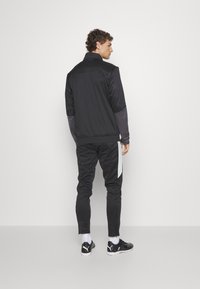 Schwarze Sportjacke mit hohem Kragen und strukturierten Ärmeln. Schwarze Hose mit einem kontrastierenden hellgrauen Streifen. Schwarze Sneakers runden das Outfit ab.