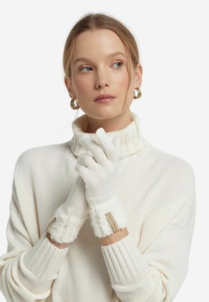 Hvide strikhandsker med en fluffy manchet og et sandfarvet mærke, båret af en model i en hvid turtleneck sweater, med hænderne samlet.