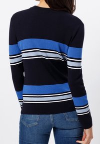 Woman wearing a long-sleeve navy sweater with blue, light blue, white, and black horizontal stripes, paired with blue denim jeans.