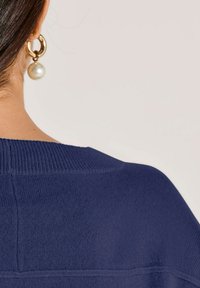 Dunkelblauer Strickpullover mit einem gerippten Ausschnitt. Verfügt über sichtbare Nähte; kombiniert mit großen goldenen Creolen und einem Perlenakzent.