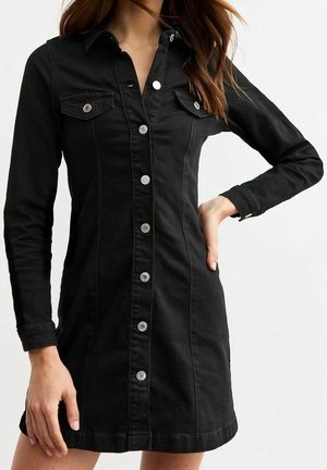 Femme portant une robe en jean noire cintrée avec des boutons, manches longues et poches à rabat sur la poitrine, debout avec une main sur la hanche.