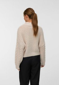 Femme avec une queue de cheval brune portant un pull en tricot épais beige avec des manches en tricot torsadé et un pantalon noir, de dos contre un fond uni.