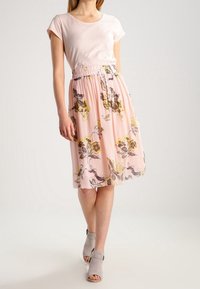 Haut rose clair à manches courtes associé à une jupe rose à motif floral avec des détails jaunes et gris ; mannequin avec des sandales à talons gris.