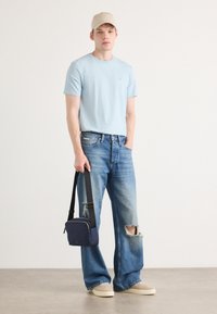 Hellblaues Baumwoll-T-Shirt, abgenutzte hellblaue Jeans, beigefarbenes Cap, hält eine kleine marineblaue Umhängetasche, cremefarbene Slipper, drinnen.