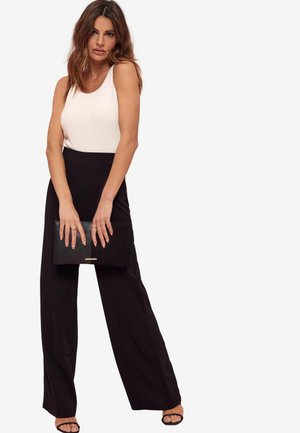 Pantaloni neri a gamba larga con una trama liscia, abbinati a un top a canotta aderente di colore rosa chiaro. Tiene una pochette nera con un accento dorato.