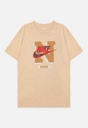 Beżowy t-shirt z krótkim rękawem z dużą beżową literą "N", czerwonym logo Nike, wzorem swoosh i napisem "SWOOSH" na środku z przodu.
