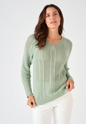 Donna con maglione a maglia aperta verde chiaro a scollo a V e pantaloni bianchi su sfondo chiaro uniforme, che tiene l'orlo del maglione.