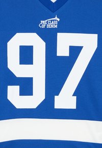 Kék mez nagy fehér "97"-es számokkal és "A DENIM OSZTÁLY" felirattal, fehér díszítésekkel és V alakú nyakkivágással.